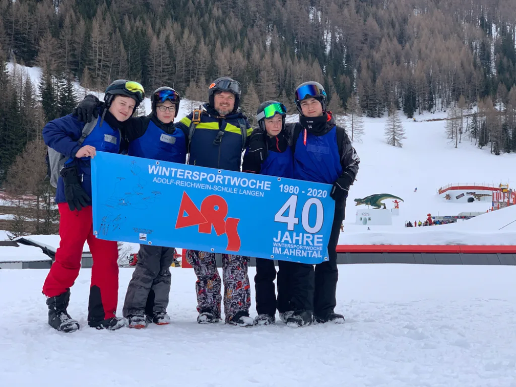 Eine Gruppe in blauen Jacken hält das Banner vor dem verschneiten Hang.